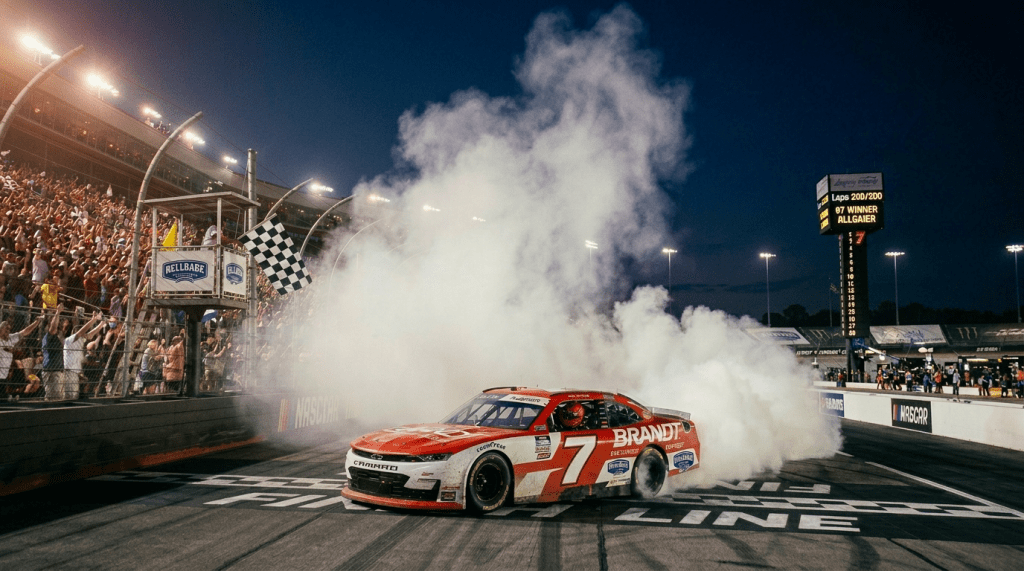 Red and white NASCAR car number 7 crossing the finish line at night with crowds cheering